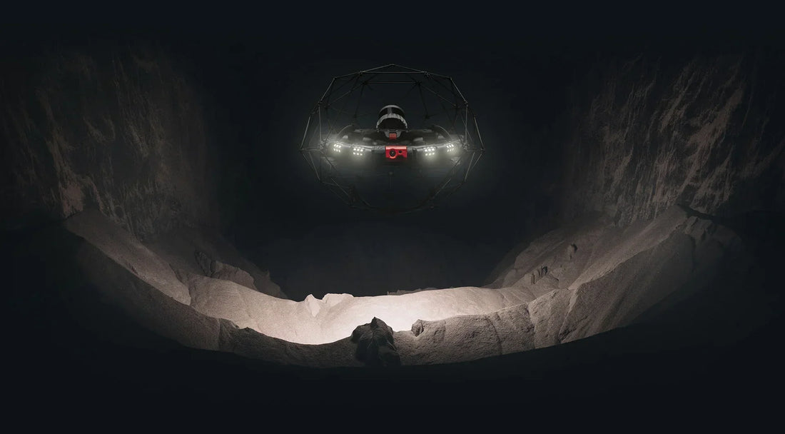 A drone equipped with bright lights hovers inside a dark, rocky cavern, illuminating the uneven ground and jagged walls below.