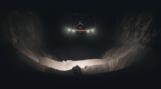 A drone equipped with bright lights hovers inside a dark, rocky cavern, illuminating the uneven ground and jagged walls below.
