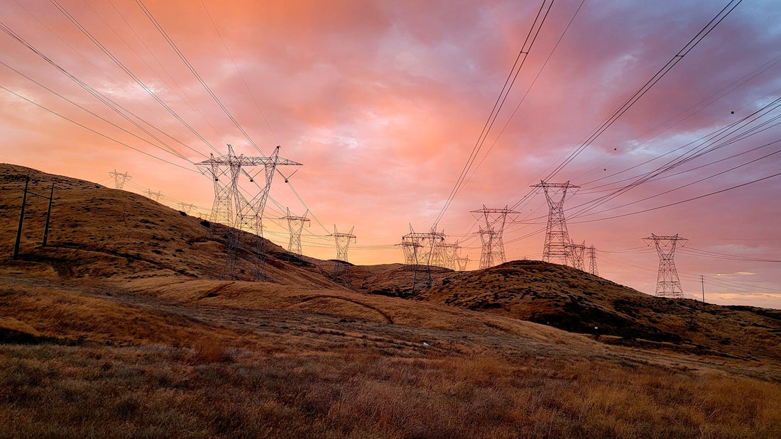 AeroVision - Wideshot at sunset, clouds of orange, pink, and purple hues in the sky. A hillside with a number electrical tower lines facing the camera.