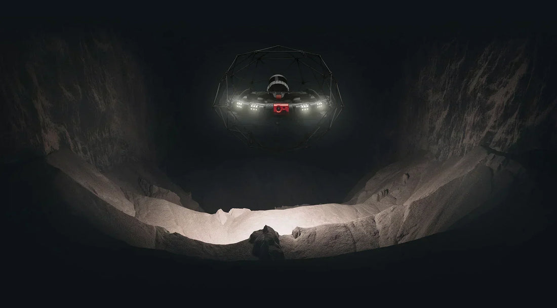 A drone equipped with bright lights hovers inside a dark, rocky cavern, illuminating the uneven ground and jagged walls below.
