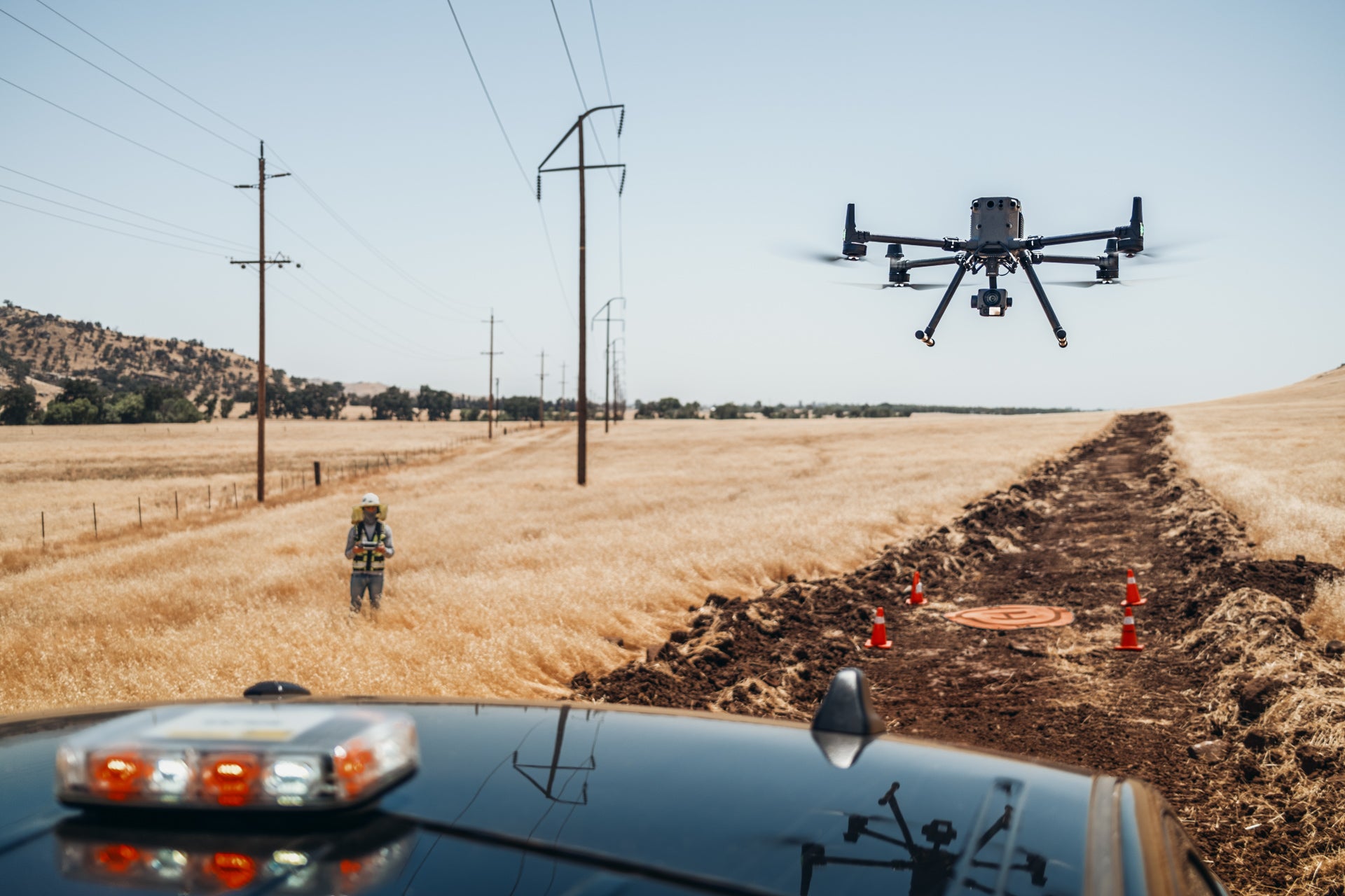 Drone flying over a field with a car in the foreground and drone pilot in the background.