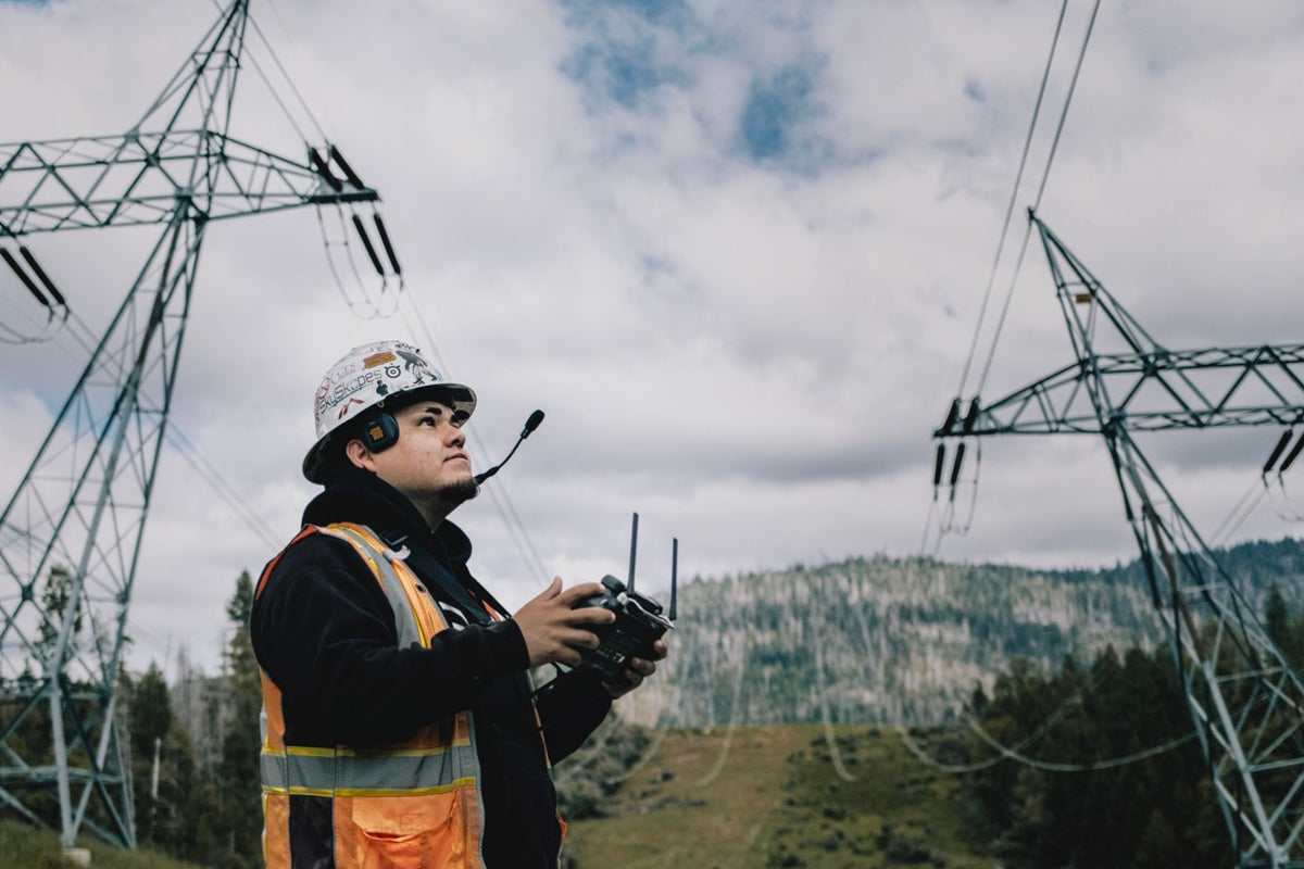 Person in safety gear operating equipment with power lines and mountains in the background
